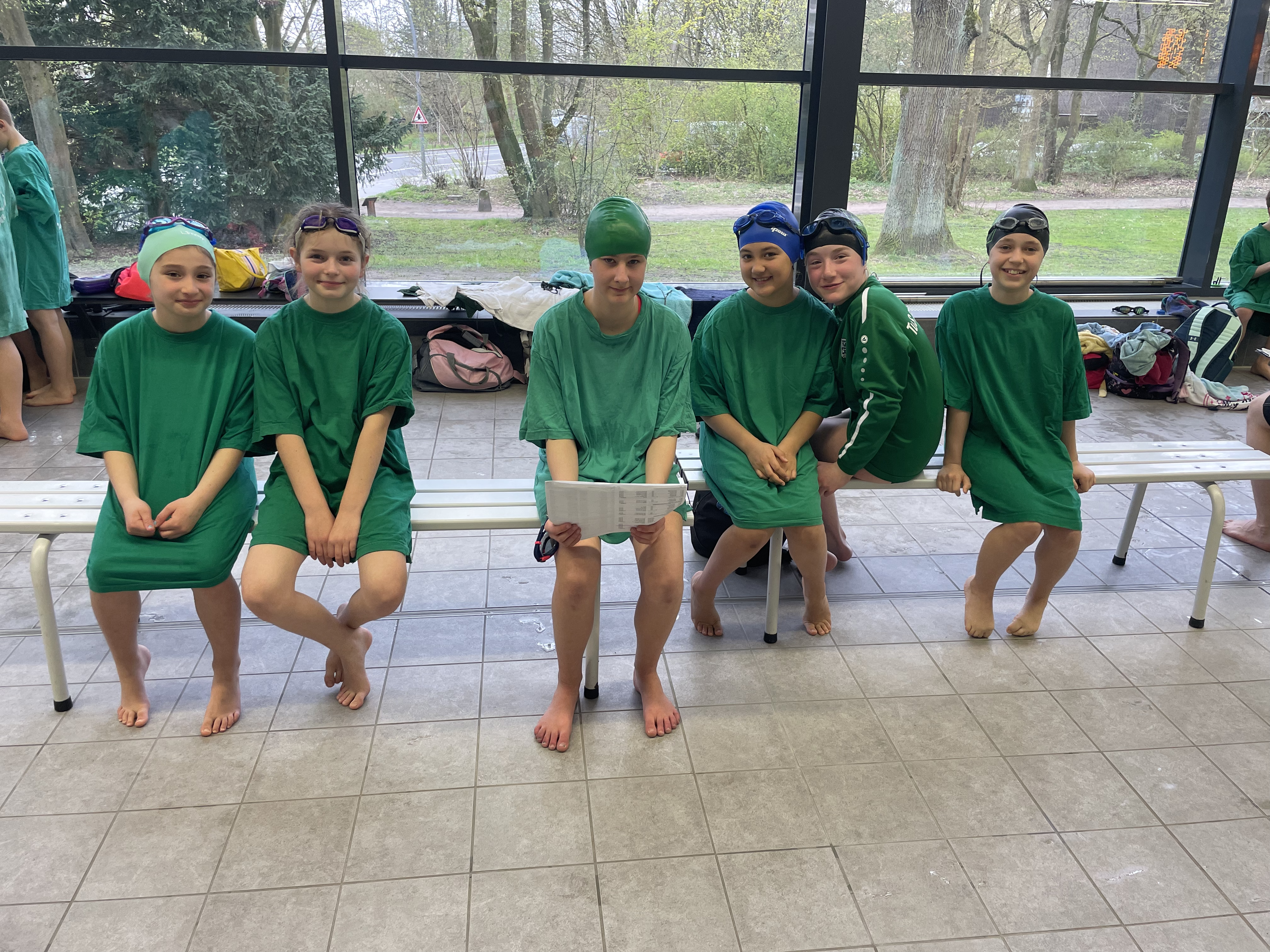 Fünf Jugendliche und Kinder sitzen in grünen T-Shirts und mit Badekappen auf in einer Schwimmhalle.