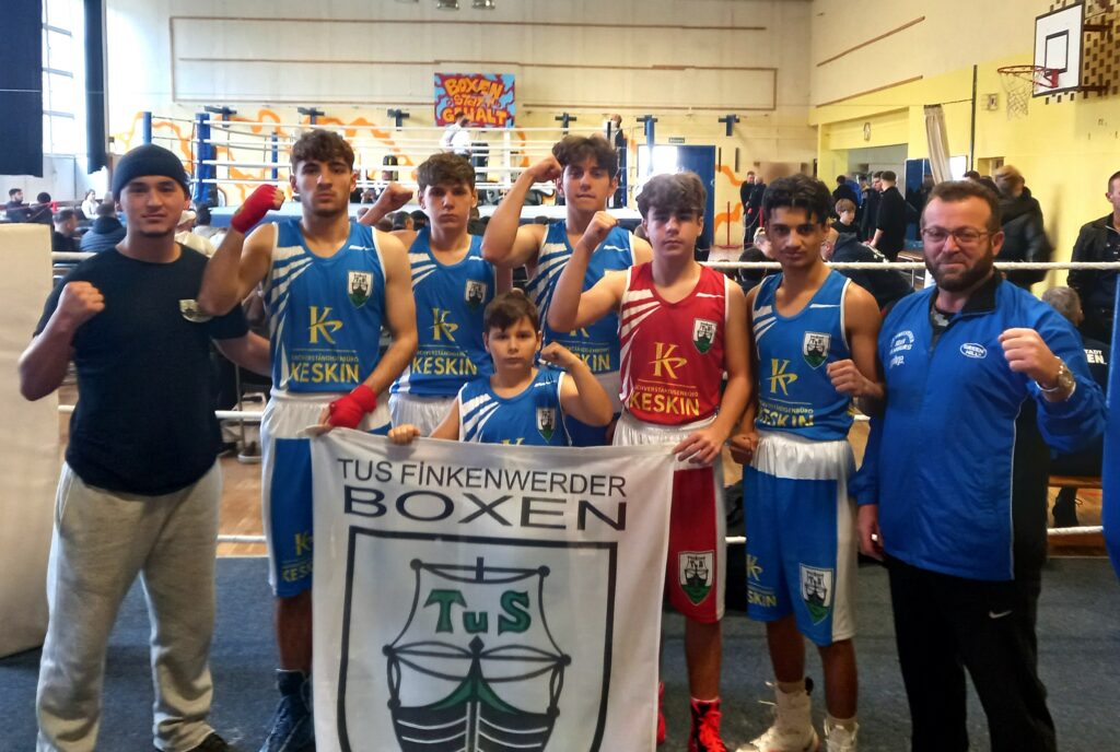 Sieben junge Boxer und der Trainer vom TuS Finkenwerder posieren im Ring, sie ballen die Faust und halten eine Flagge vom TuS Finkenwerder vor sich.