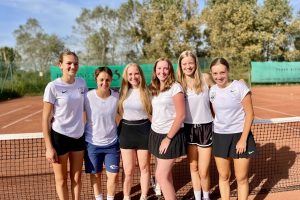 Sechs junge Frauen in weißen T-Shirts stehen auf einem Outdoor Tennisplatz. Die Sonne scheint.