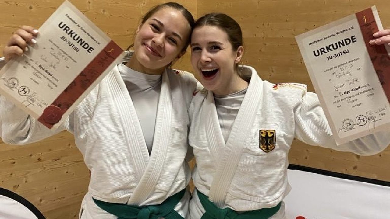 Zwei Sportlerinnen der Ju-Jutsu Abteilung halten ihre Urkunden in die Kamera und freuen sich über ihre Bestandene Prüfung. Sie haben einen weißen Anzug und einen Grünen Gürtel um.