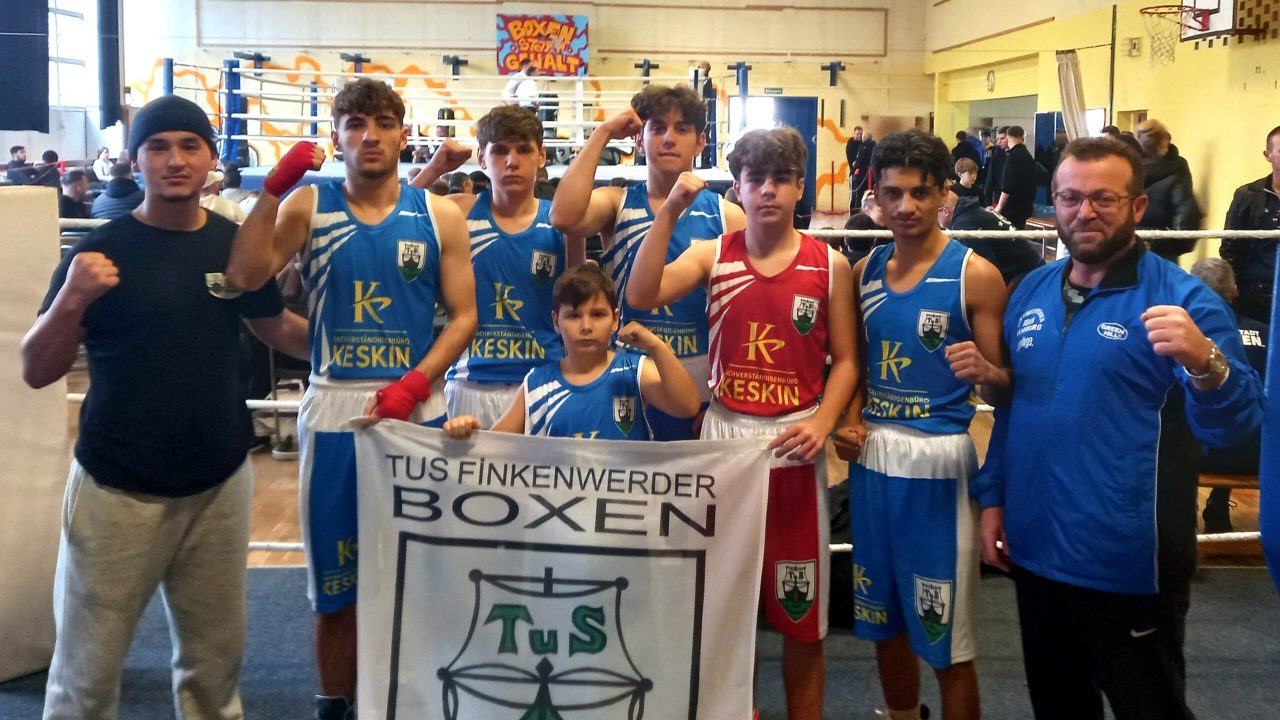 Sieben junge Boxer und der Trainer vom TuS Finkenwerder posieren im Ring, sie ballen die Faust und halten eine Flagge vom TuS Finkenwerder vor sich.