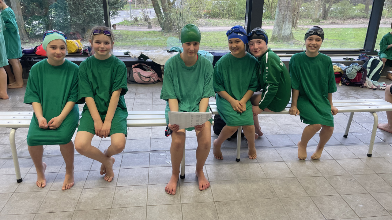 Fünf Jugendliche und Kinder sitzen in grünen T-Shirts und mit Badekappen auf in einer Schwimmhalle.